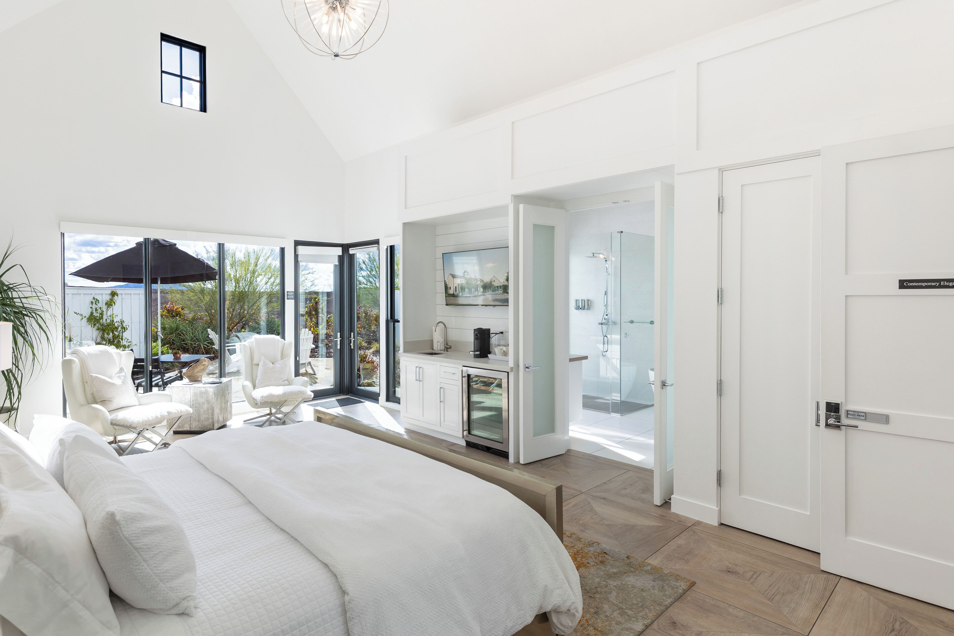 Quarto moderno com cama grande, área de estar com cadeiras, cozinha e chuveiro em box de vidro.
