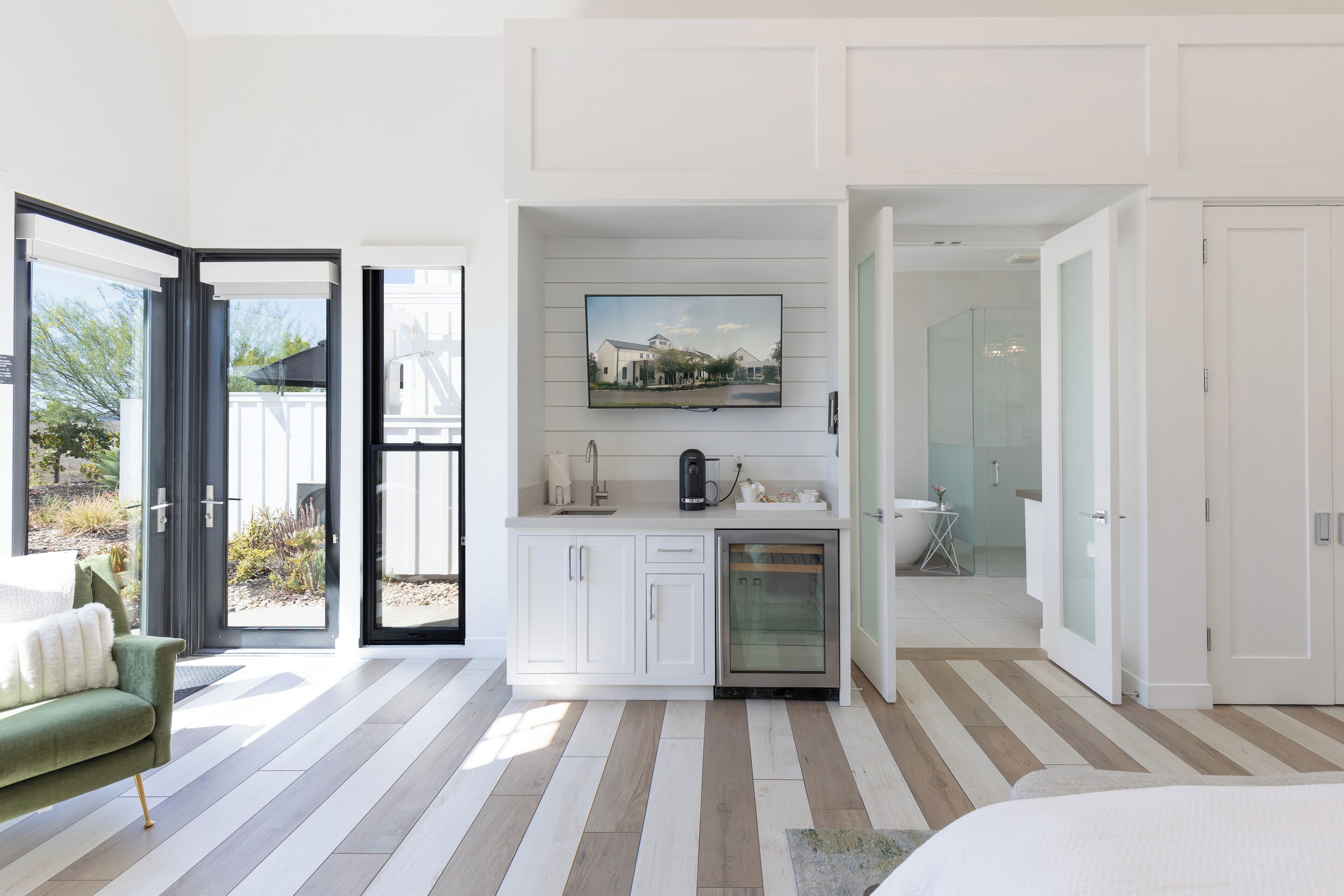 Habitación moderna y bien iluminada con una cama grande, un sillón verde, una zona de cocina con cafetera y un baño con cabina de ducha de vidrio.
