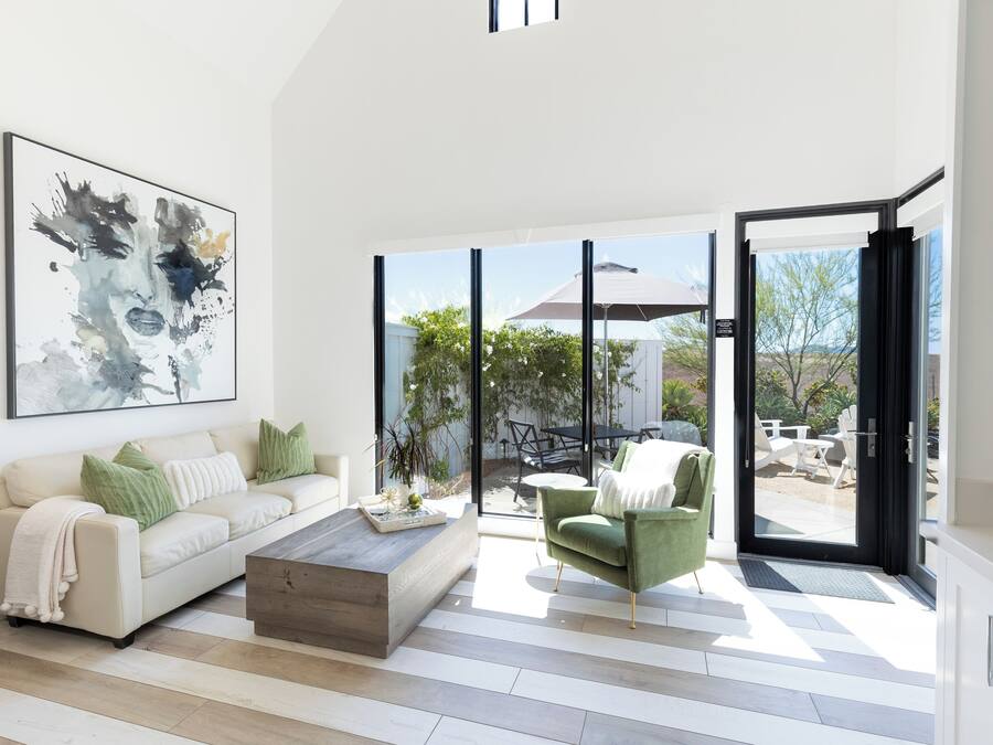 Un salon moderne avec un canapé, une table basse et un fauteuil vert. Un grand tableau abstrait orne le mur, et de grandes fenêtres offrent une vue sur l’extérieur.