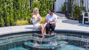 Two people sitting by a pool with a tiled edge and a white chair nearby. - The New Inn (Temecula)
