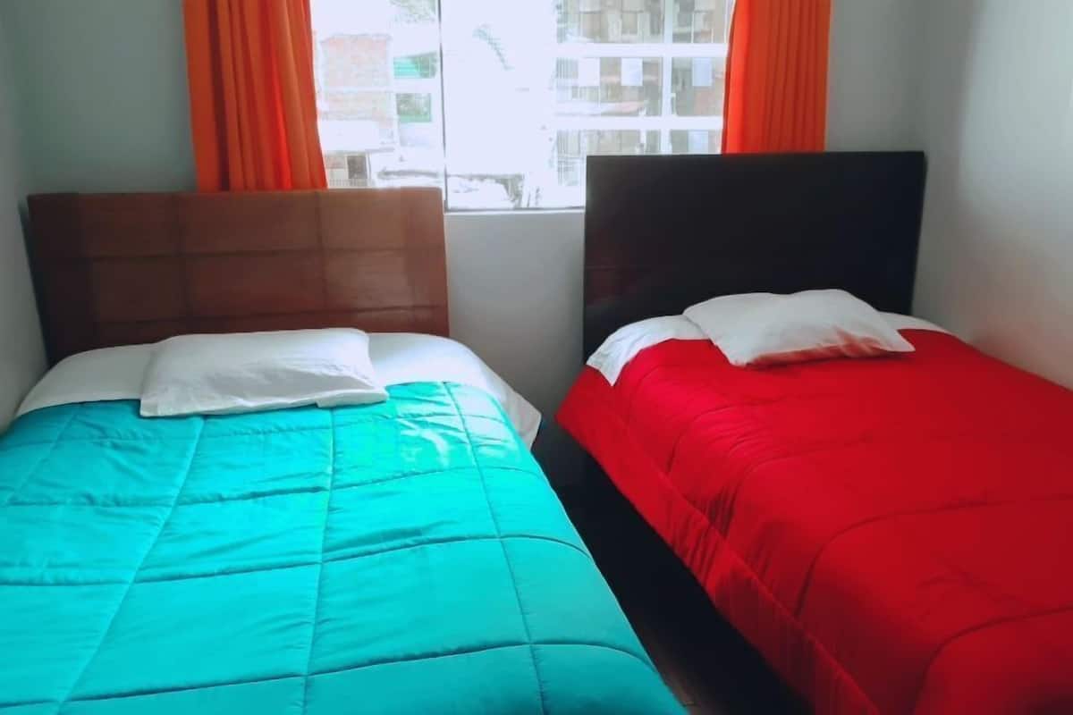 Two single beds with headboards, one with a teal bedspread and the other with a red bedspread, in a room with orange curtains and a window view.
