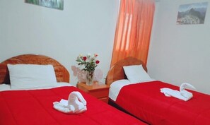 A hotel room with two beds, red bedding, white pillows, a small table with a vase of flowers, and a view of mountains through the window. - Modo Hostel Resto Bar (Machu Picchu)