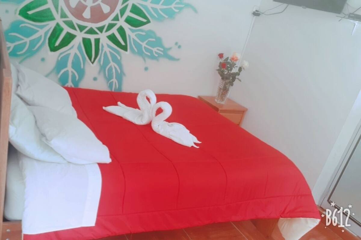 A neatly made bed with a red bedspread and white pillows, a decorative wall mural, and a vase with flowers on a bedside table.