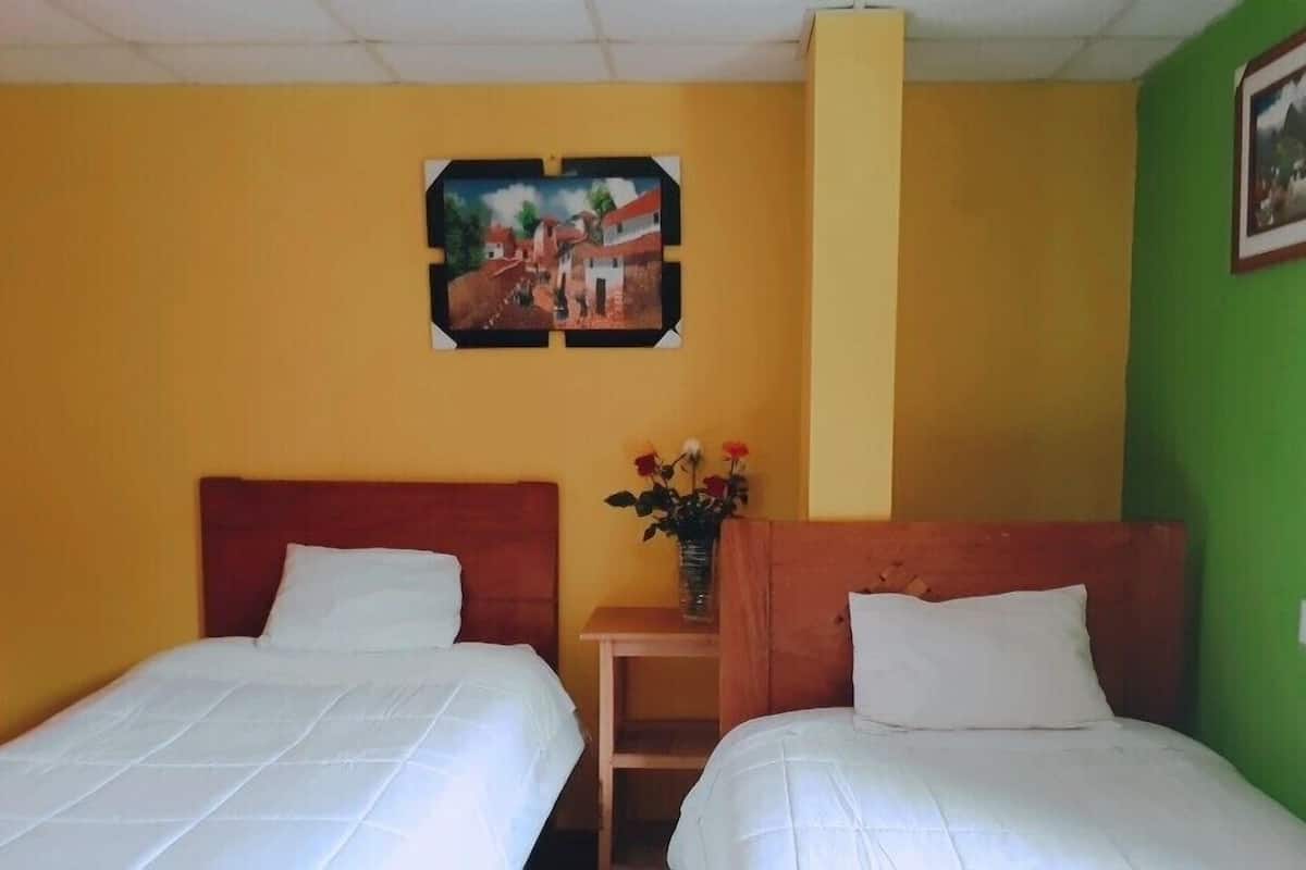 A hotel room with two single beds, a small table with a vase of flowers, and a painting on the wall.