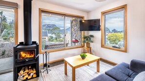 Un salon moderne doté d’un poêle à bois, de grandes fenêtres offrant une vue sur un paysage montagneux, d’un téléviseur à écran plat fixé au mur et d’un coin salon confortable avec un canapé et une petite table.