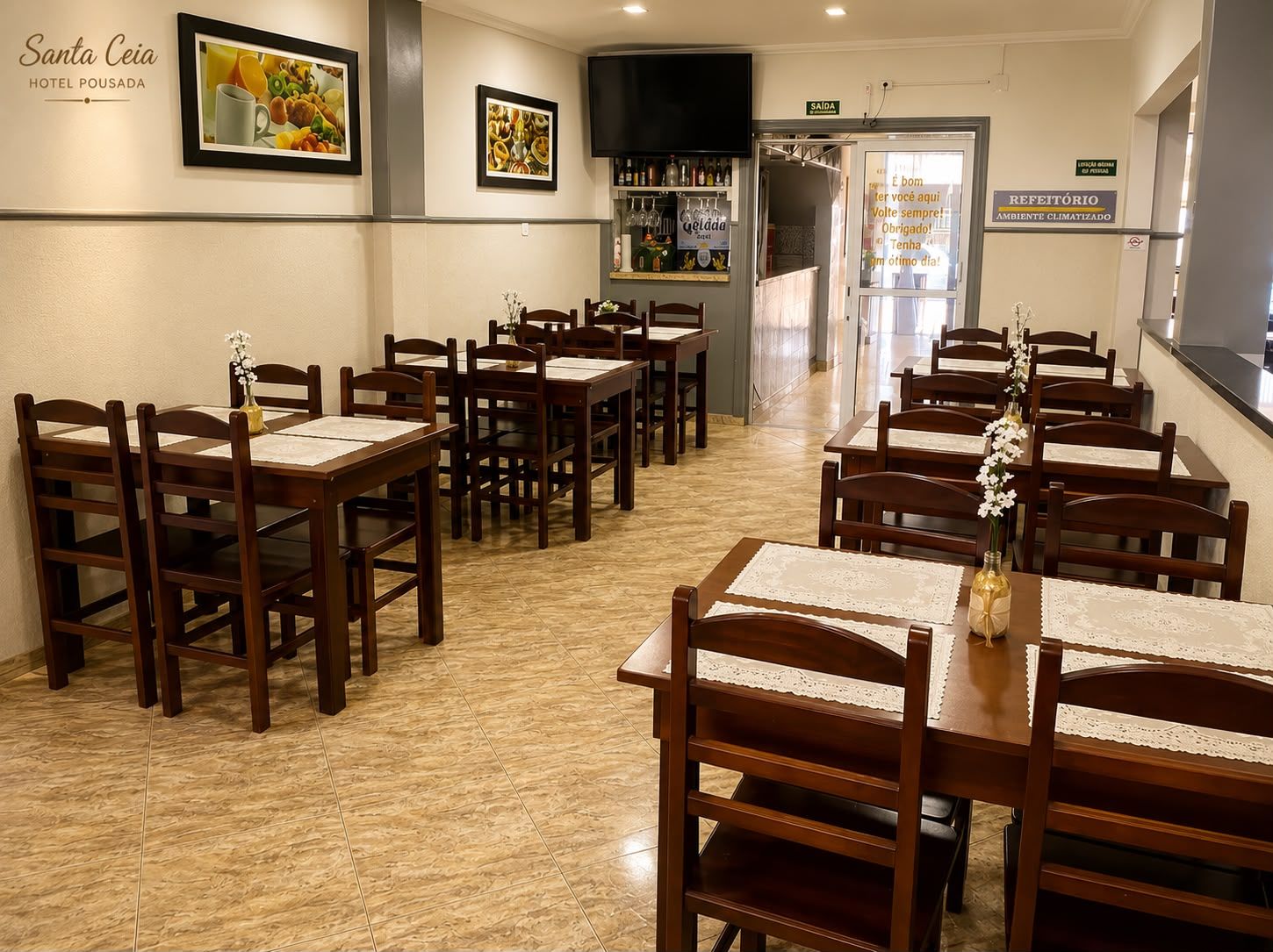 A hotel dining area with multiple wooden tables and chairs, a bar area with bottles, and framed pictures on the walls.