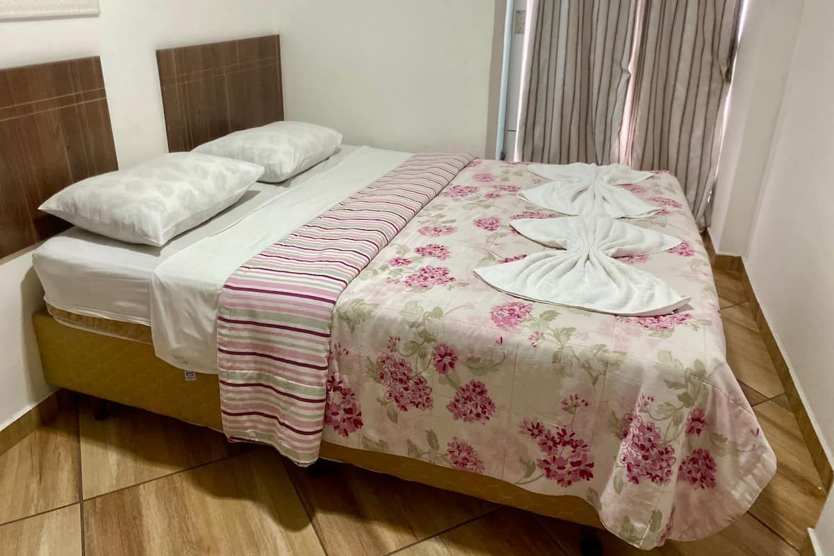 A neatly made bed with a floral bedspread and striped blanket, a headboard, and a window with vertical blinds.