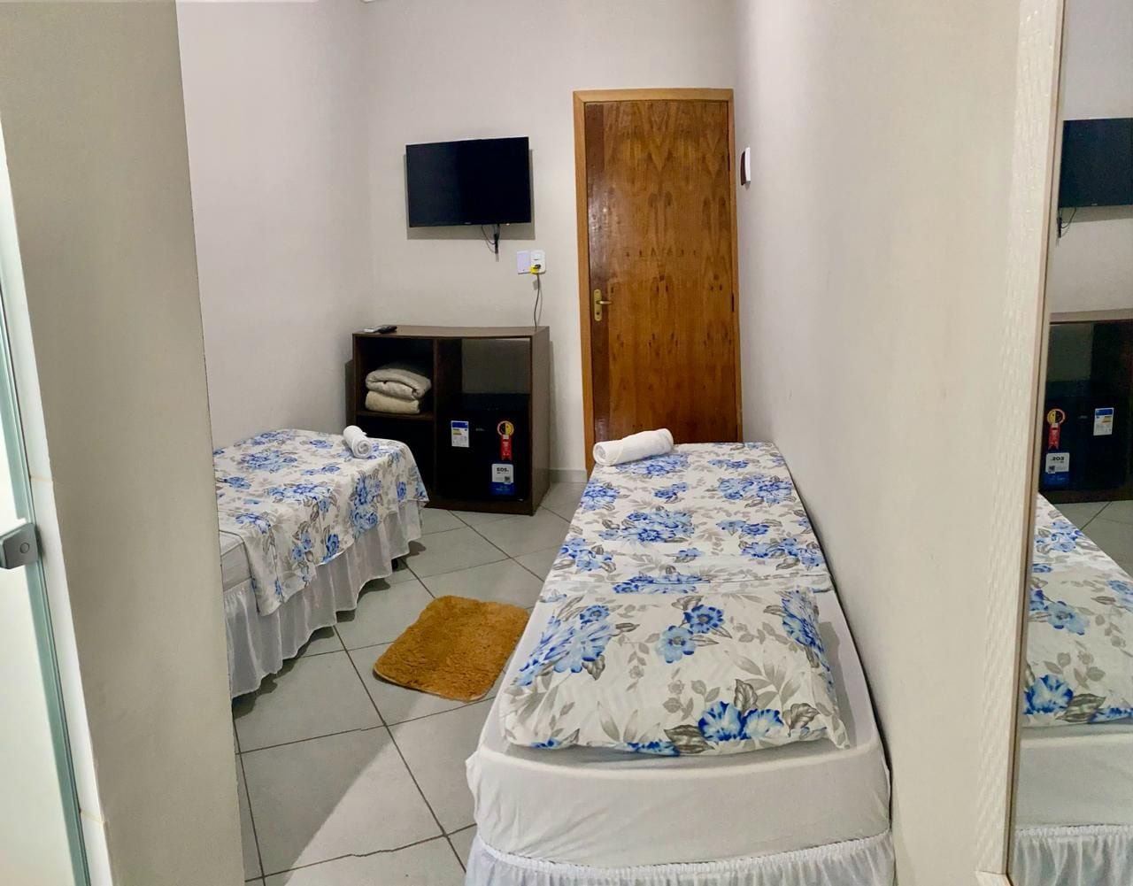 A hotel room with two single beds, a wooden door, a television on the wall, and a small desk with a chair.
