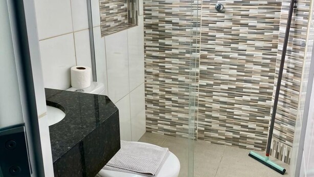 A bathroom with a toilet, a shower with a glass door, and a tiled wall. - Hotel Pousada Santa Ceia (Aparecida)