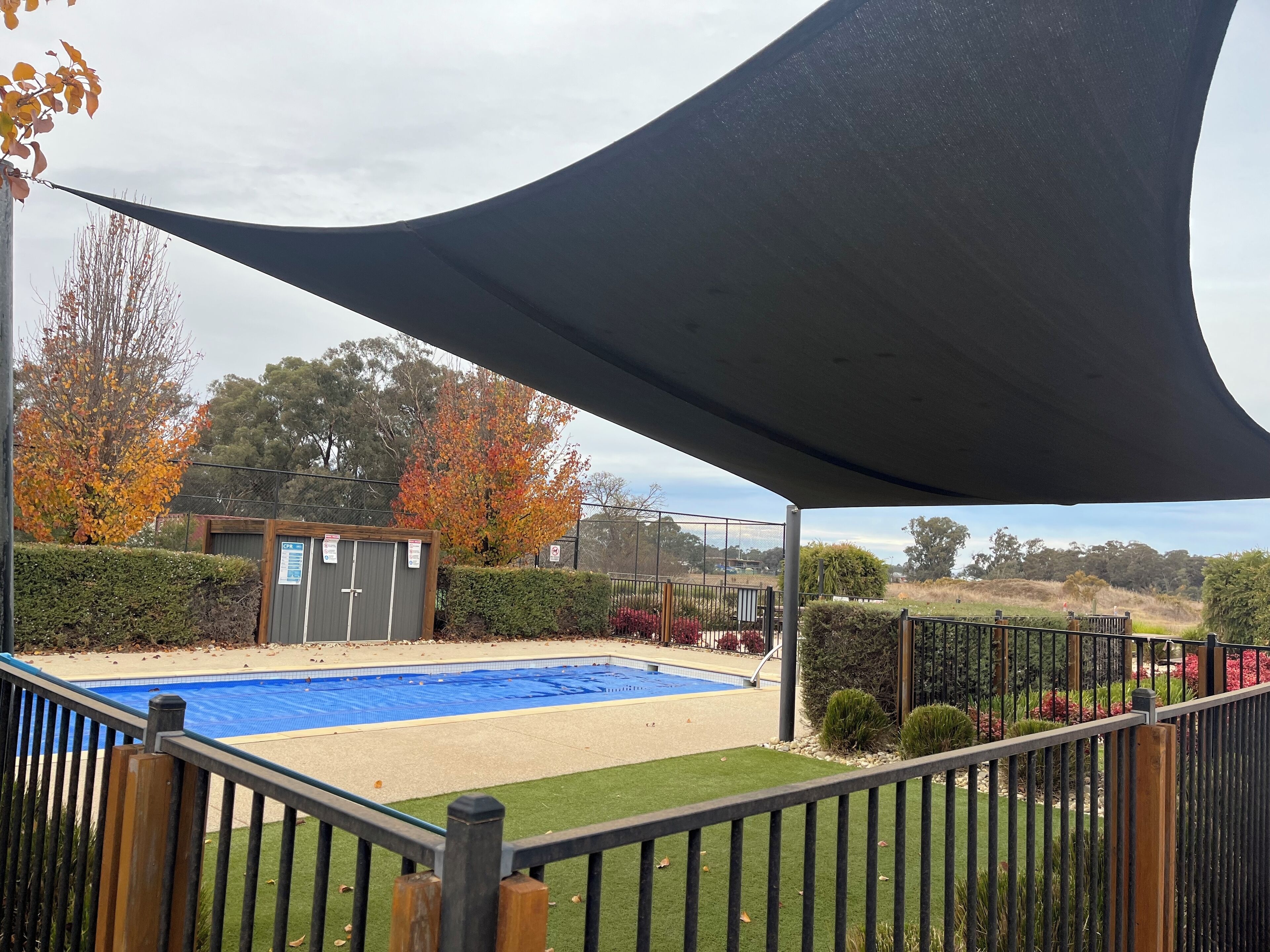 A swimming pool with a surrounding deck, a shaded area, and a fenced garden with various plants and trees.