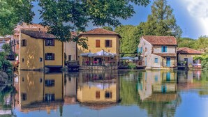 Une rivière paisible où se reflètent des bâtiments à l’architecture traditionnelle, un espace extérieur abrité avec des sièges, et un ciel dégagé.
