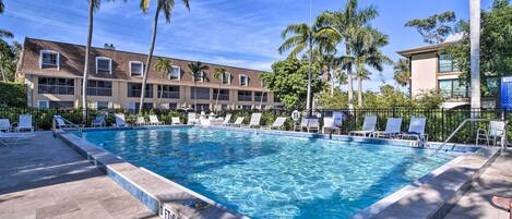 A rectangular swimming pool with clear blue water, surrounded by white lounge chairs and palm trees.
