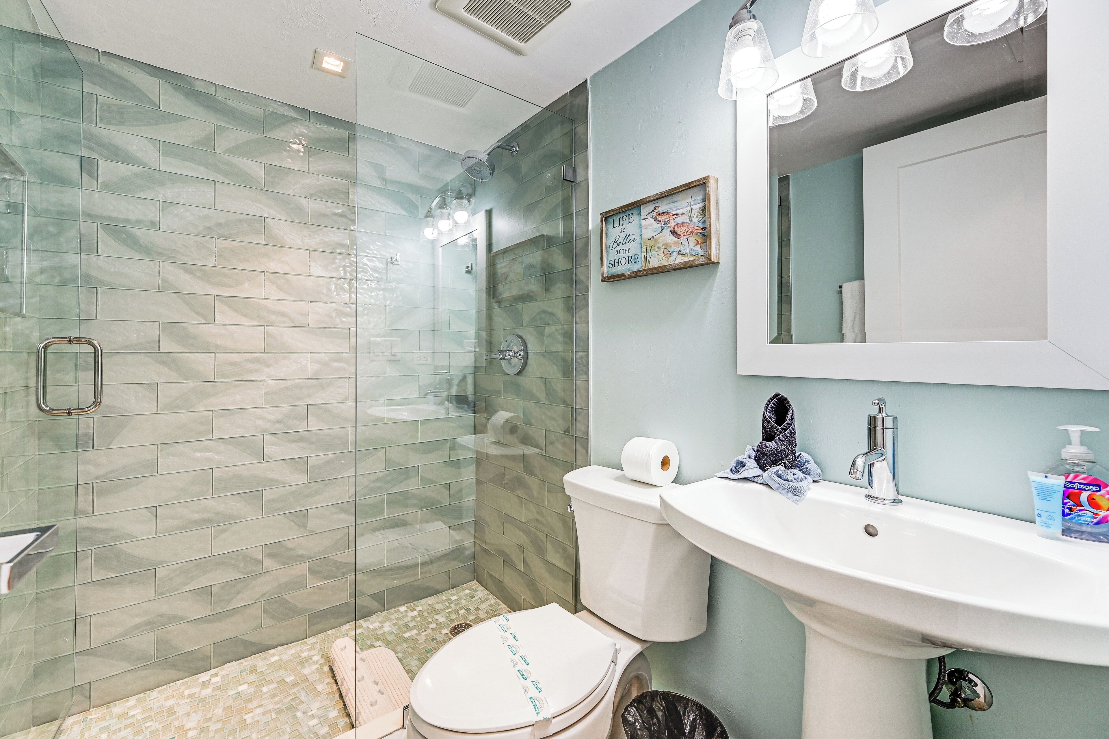 A bathroom with a glass shower enclosure, a toilet, a sink with a mirror above, and a framed picture on the wall.