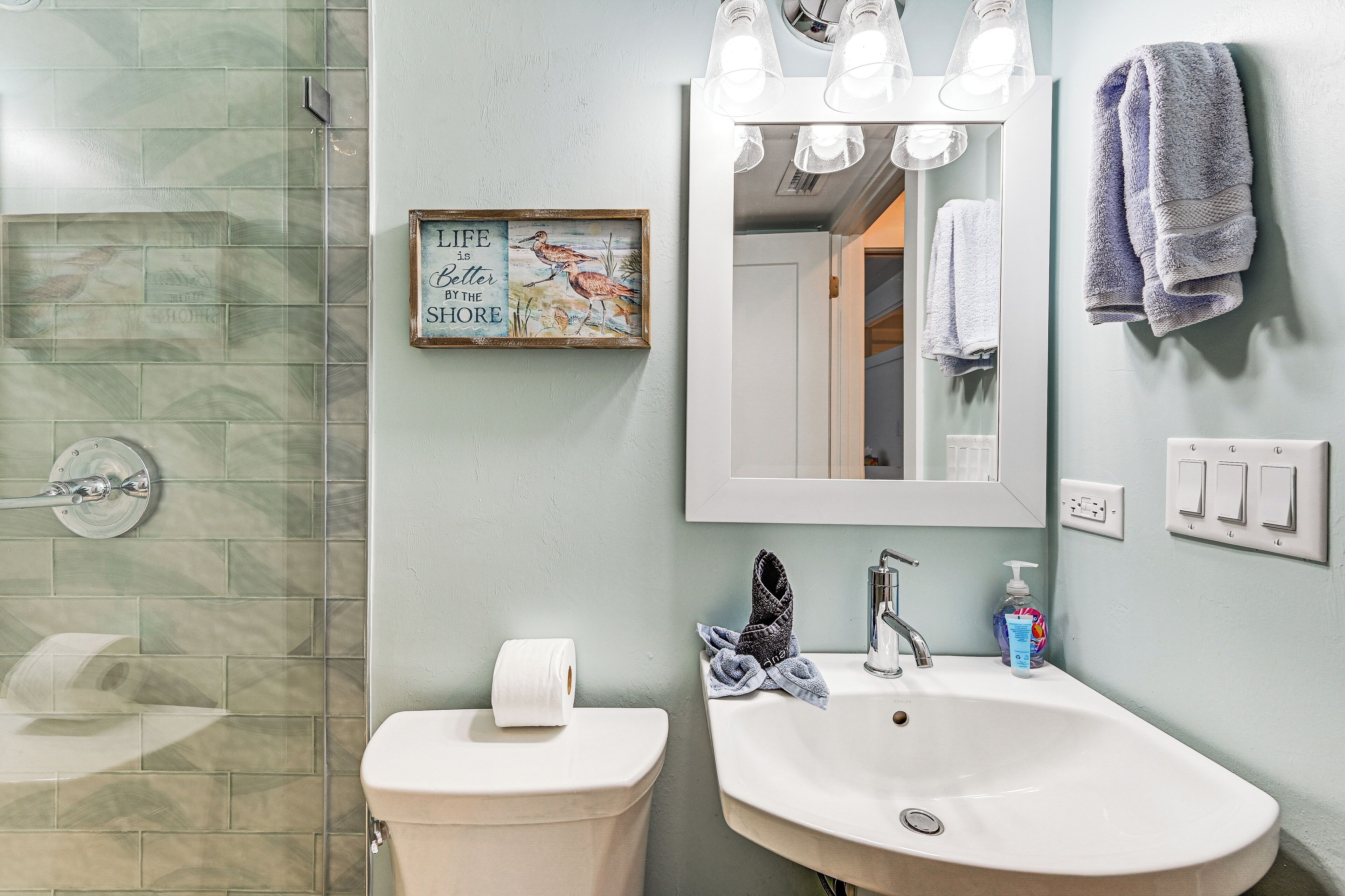 A bathroom with a shower, toilet, sink, and a mirror with lights. There's a framed quote on the wall and a towel hanging on the wall.