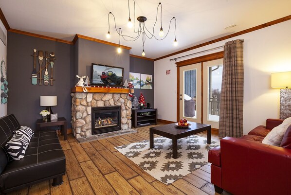 A living room with a stone fireplace, a black leather sofa, a red sofa, a wooden coffee table, and a chandelier. - Luxury 3 Bedroom Blue Chalet. Entire House. 2 minute drive from the village (The Blue Mountains)