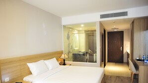 Habitación de hotel con una cama grande, un escritorio, una silla y un baño visible a través de una puerta de vidrio.