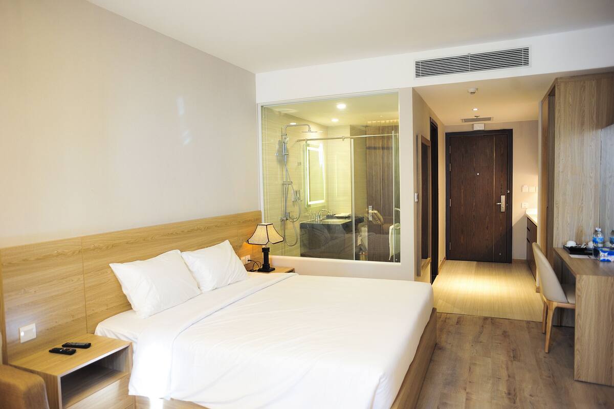 A hotel room with a large bed, a desk, a chair, and a bathroom visible through a glass door.