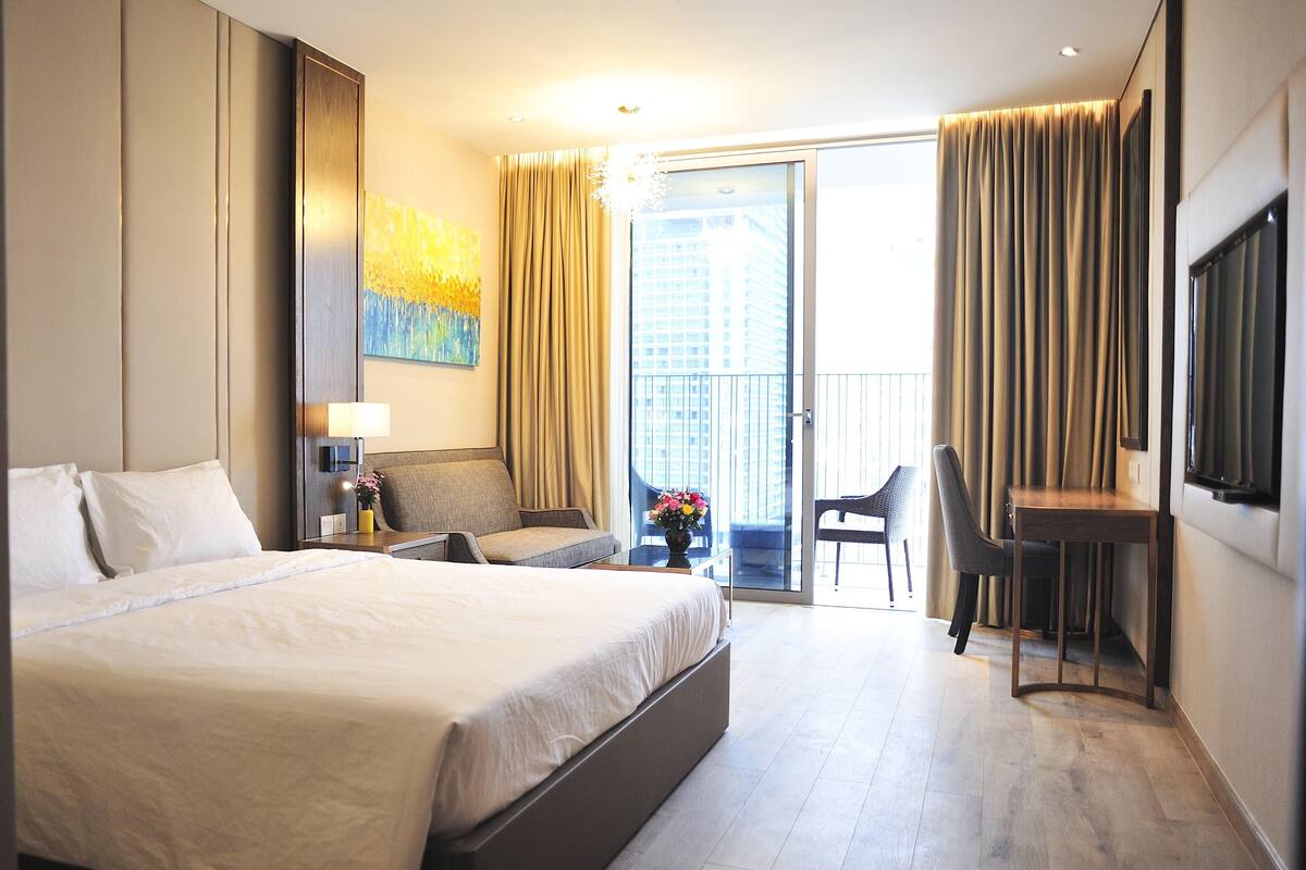 A hotel room with a large bed, a desk, a chair, a TV, and a balcony with curtains.
