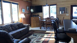 A cozy living room with a sofa, a dining table with chairs, a TV, and a large window with a view of trees.