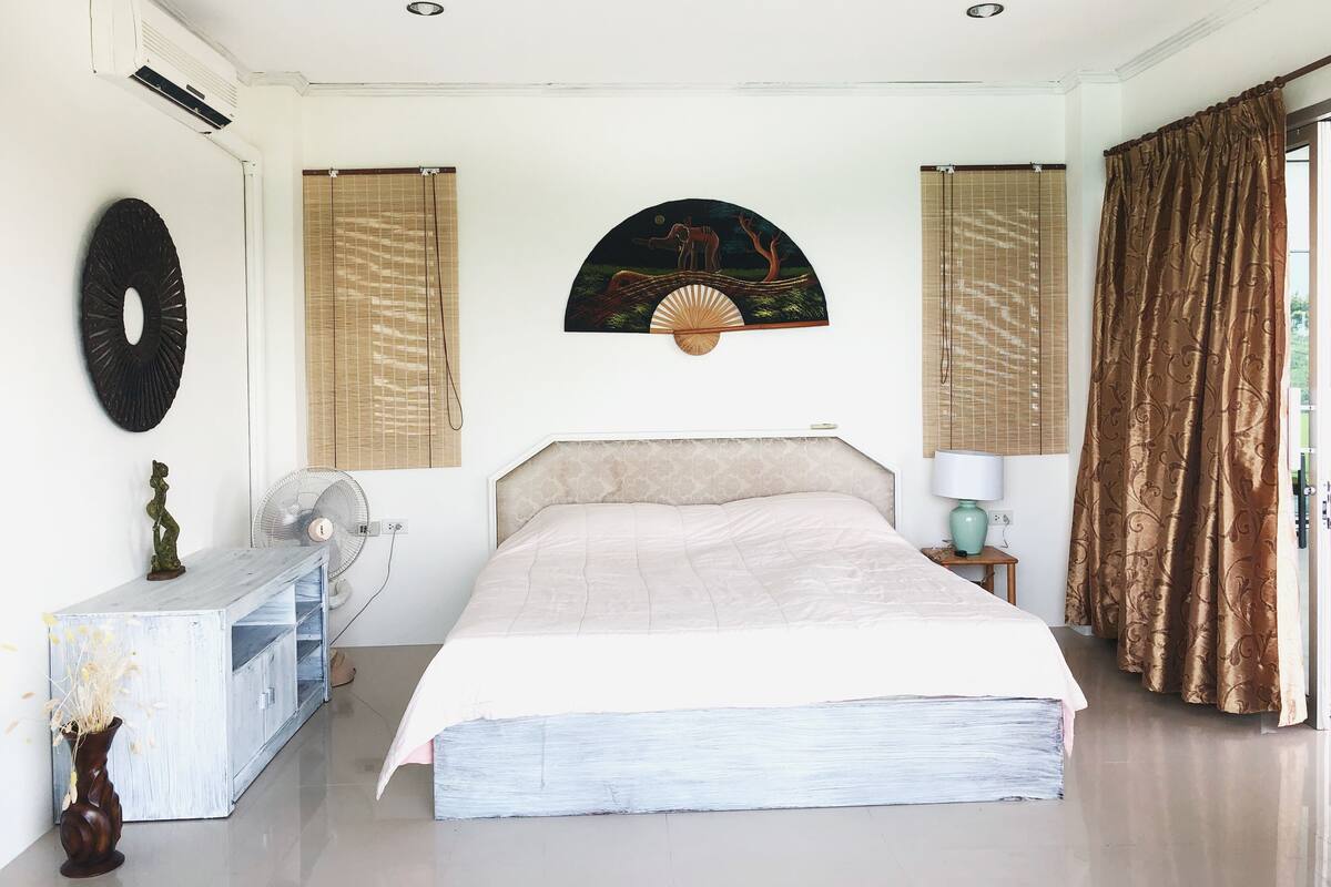 A bedroom with a bed, a fan, a decorative wall piece, and a window with curtains.
