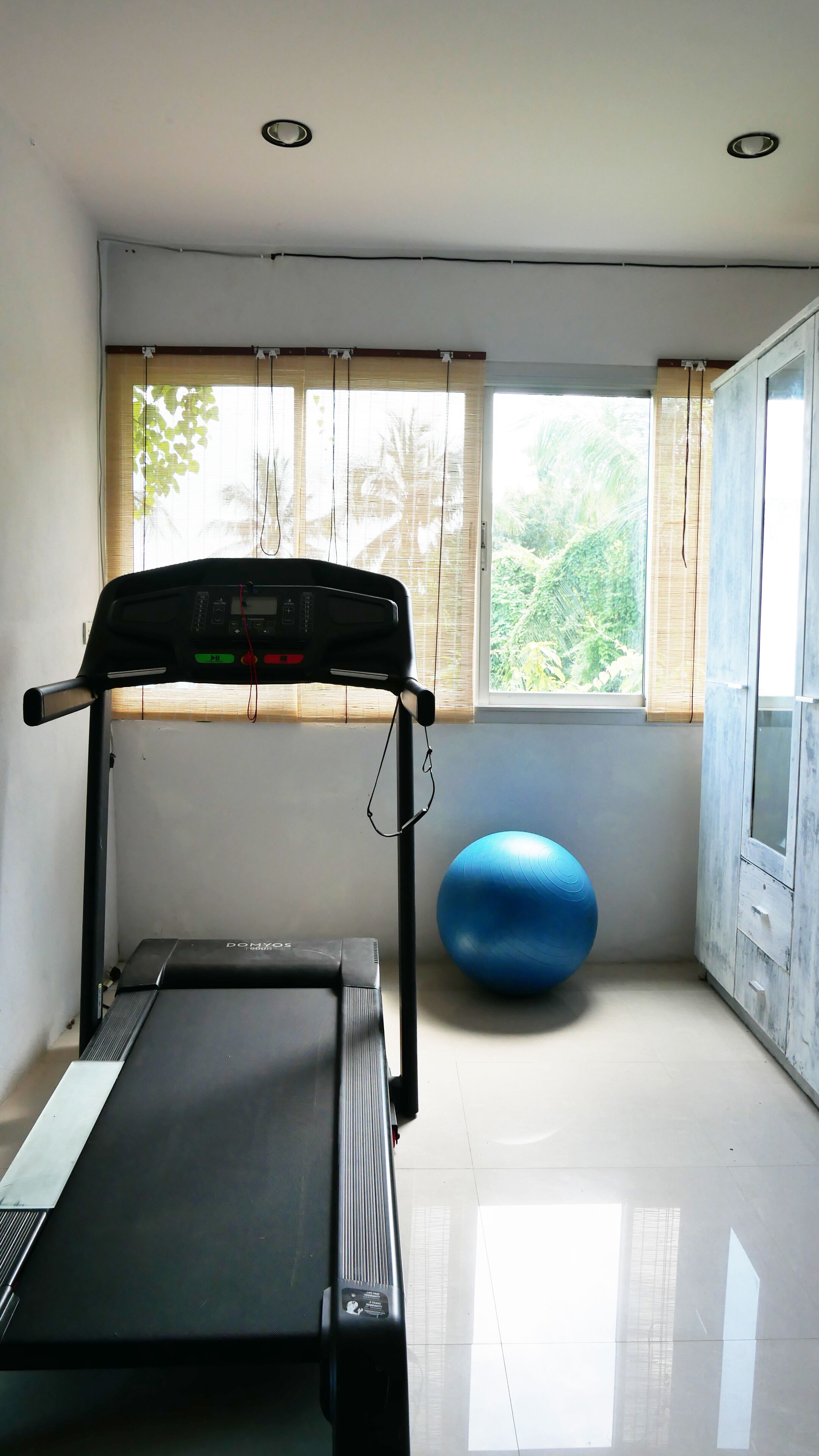 A room with a treadmill, a blue exercise ball, and a window with blinds.
