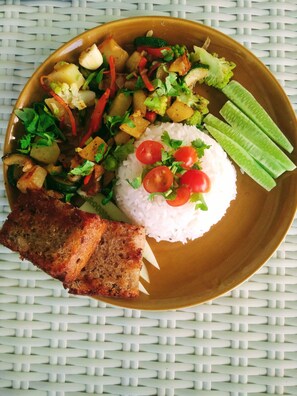 A plate with rice, vegetables, and a piece of meat on a woven tablecloth. - Your Home Samui (Koh Samui)
