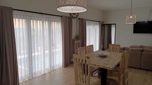 A dining area with a wooden table and chairs, large windows with sheer curtains, a television mounted on the wall, and a sofa in the background. - Private Villa with pool. Top location, a short walk to the beach (Canggu)