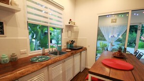Una cocina con encimera de madera, un fregadero y una ventana con persianas. Se aprecia una mesa de comedor con un mantel individual rojo y un jarrón encima.