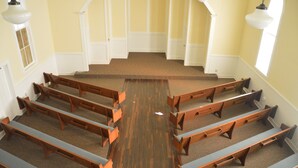 Interno di una chiesa con panche di legno disposte a file, pavimenti rivestiti di moquette e pareti bianche con una parete gialla a contrasto.