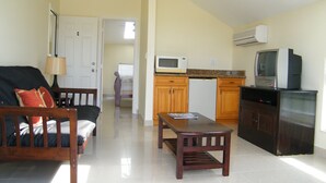 A compact living space with a sofa, a small dining table, a kitchen area with a microwave and a refrigerator, and a television on a cabinet. - Comfy Villa Suites in Long Bay Beach (Long Bay Hills)