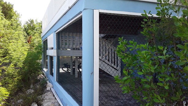 A blue building with a covered outdoor area, a chain-link fence, and some greenery. - Comfy Villa Suites in Long Bay Beach (Long Bay Hills)
