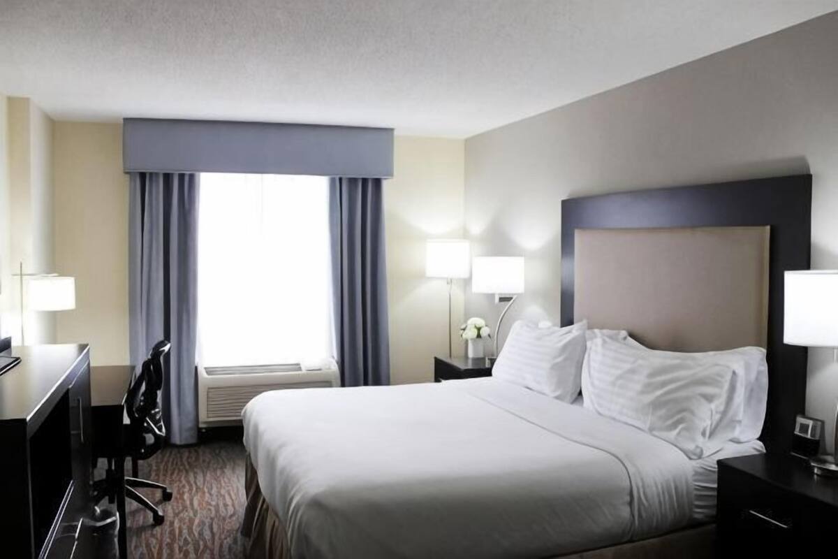A hotel room with a large bed, a desk, a chair, a nightstand with a lamp, and a window with curtains.
