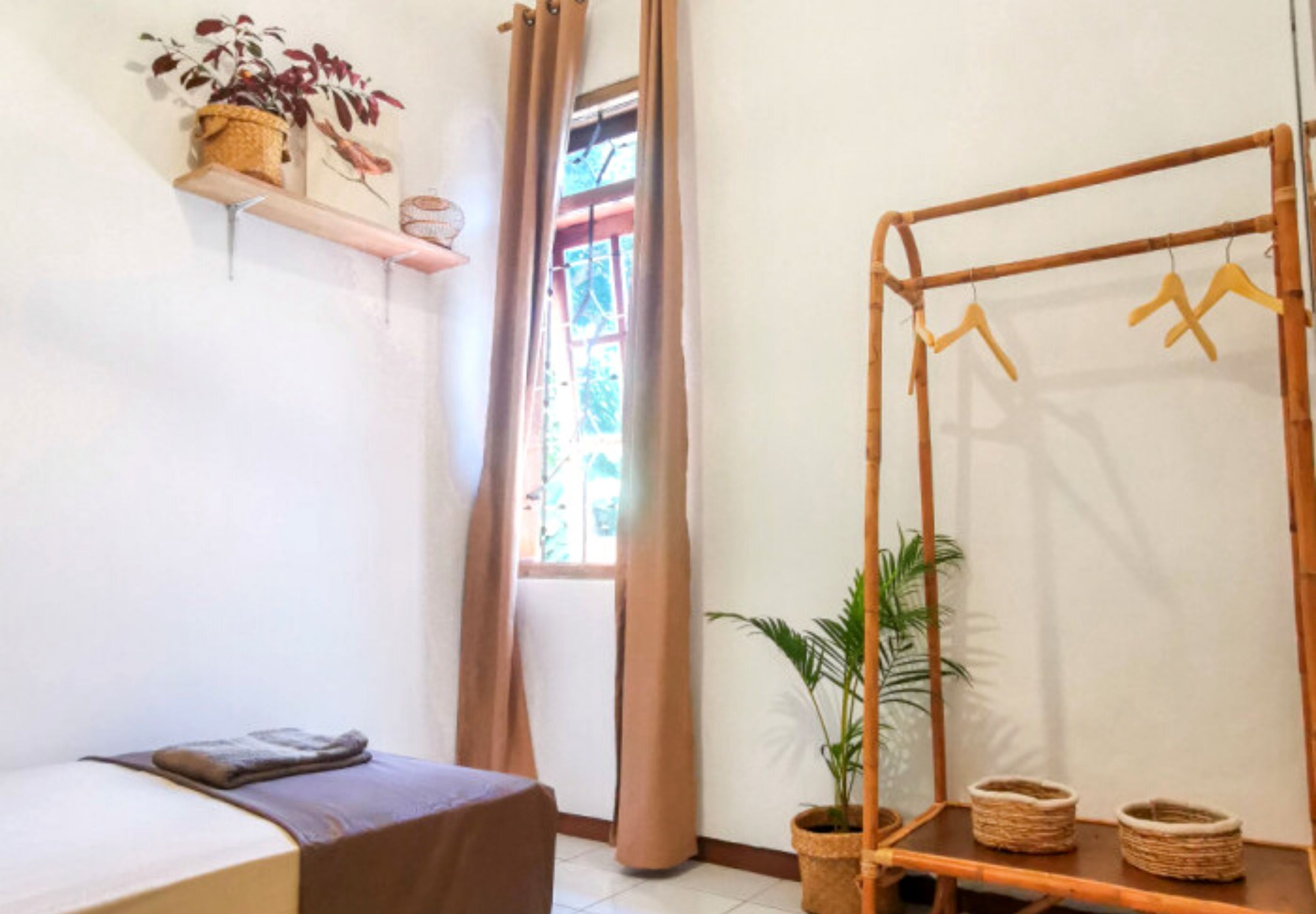 A room with a bed, a hanging clothes rack, a window with curtains, and a shelf with plants.