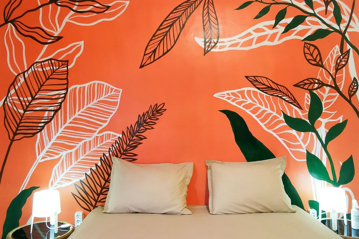 A bed with white bedding in a room with an orange wall featuring large leaf patterns.