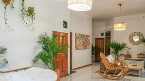 Un salón amplio con una butaca grande y acolchada, un sofá de mimbre y una mesa de comedor. Se observan varias plantas en macetas y un colgador de luz.