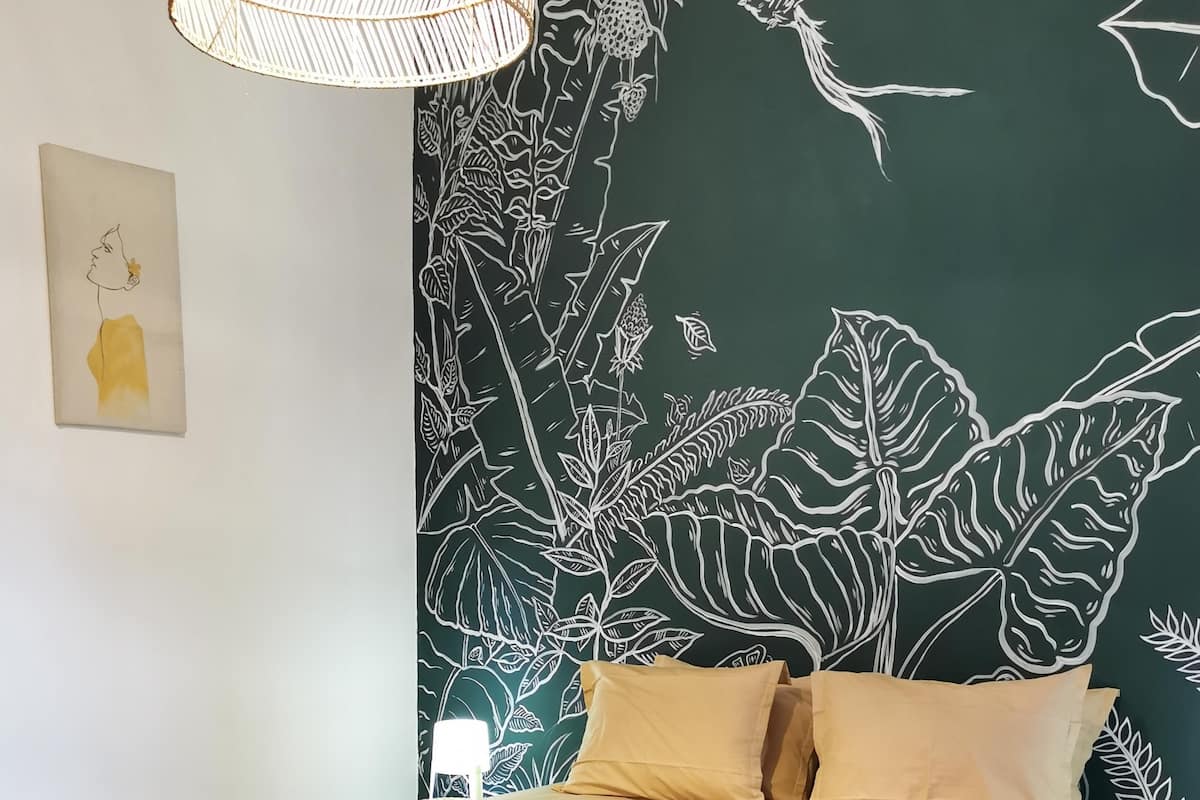 A bedroom with a bed, pillows, a wall mural, a hanging light fixture, and a small table with a lamp.