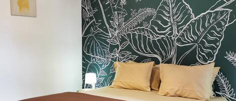 Un dormitorio con cama, almohadas, un mural en la pared, una lámpara colgante y una mesita de noche con lámpara.