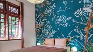 Un dormitorio con cama, ventana con cortinas, pared con estampado floral y mesita de noche con lámpara.