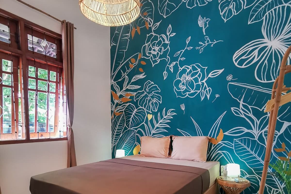 A bedroom with a bed, a window with curtains, a wall with a floral pattern, and a bedside table with a lamp.