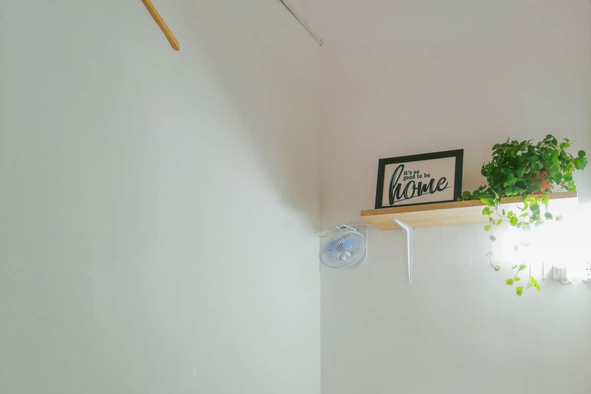 A bedroom with a bed, a wall-mounted shelf with a plant, a wall fan, and a sign that reads "it's so good to be home."
