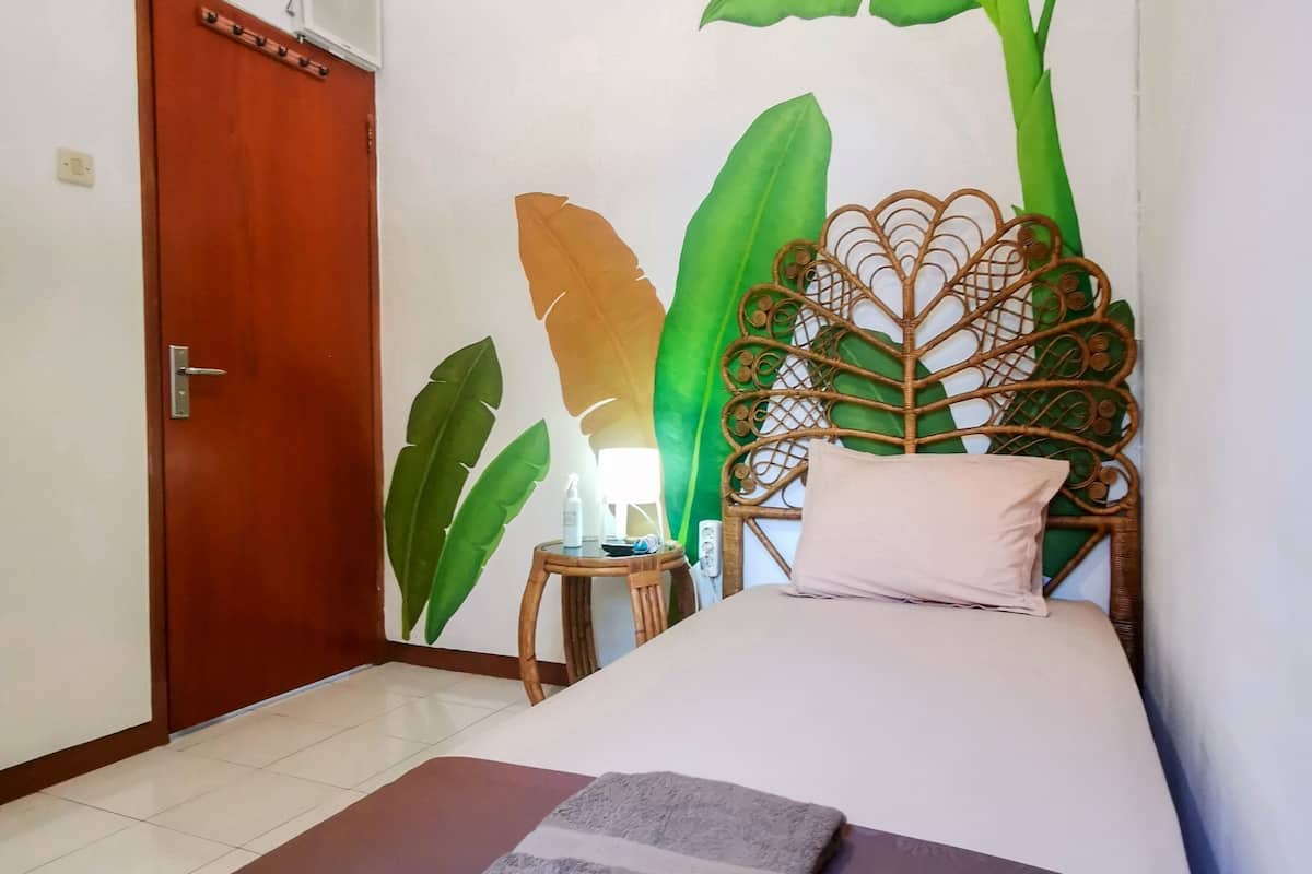 A room with a bed, a wooden door, a wall mural, a small table with a lamp, and a folded towel on the floor.