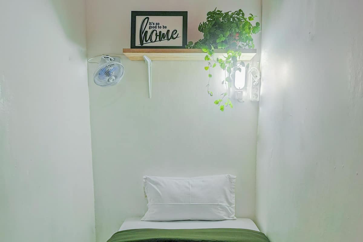 A small, single-bed room with a green bedspread, a white pillow, a brown towel, a wall-mounted fan, a framed sign saying "it's so good to be home", and a potted plant.