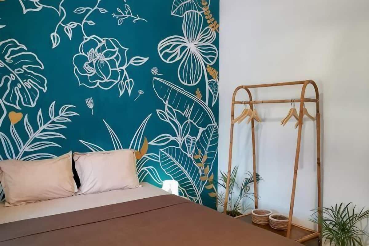 A bedroom with a bed, a wall with a floral pattern, a bamboo clothes rack, and a small plant.