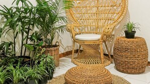 Una silla de mimbre con asiento acolchado, rodeada de plantas en macetas y un otoman tejido sobre un piso de baldosas.
