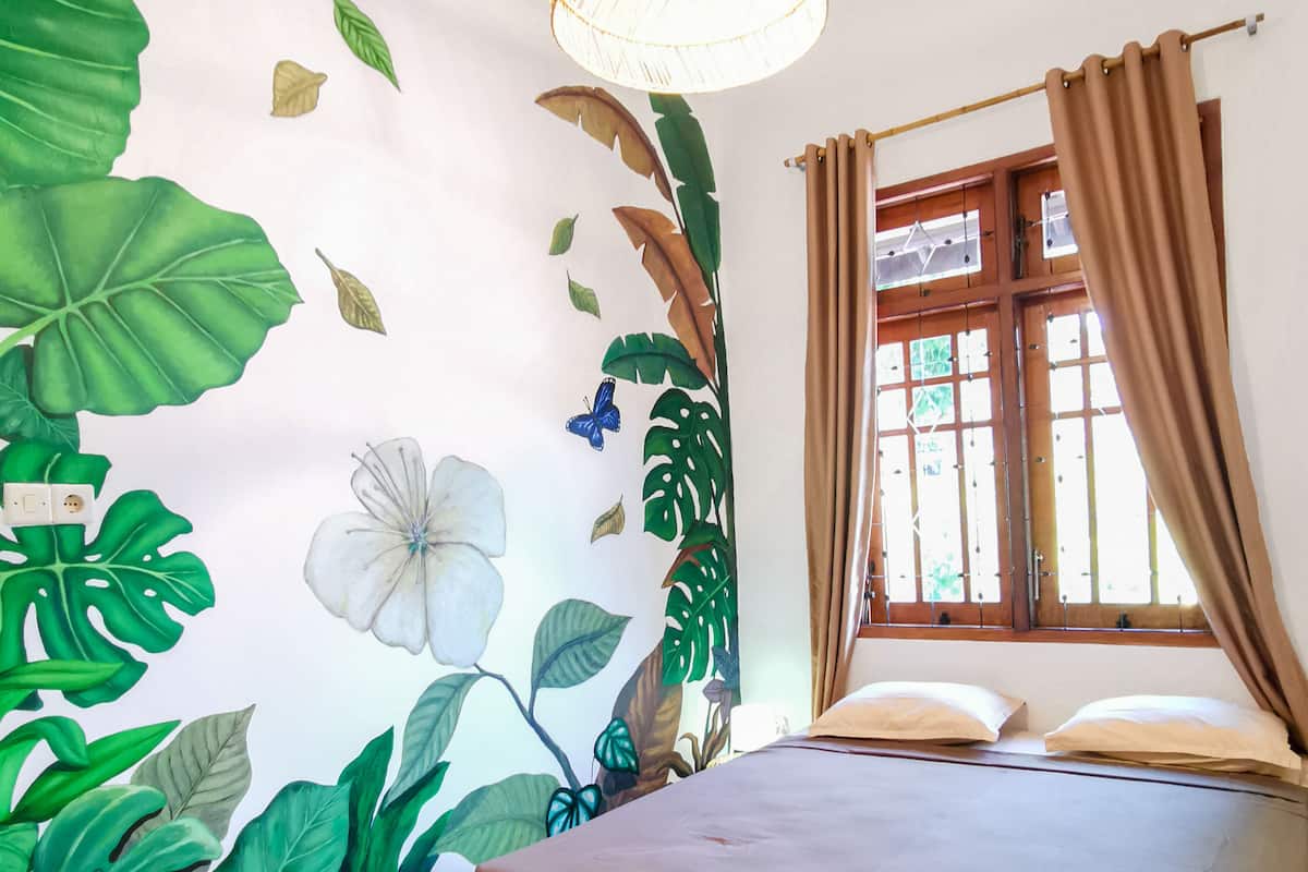 A bedroom with a bed, a window with curtains, and a wall with a tropical mural.