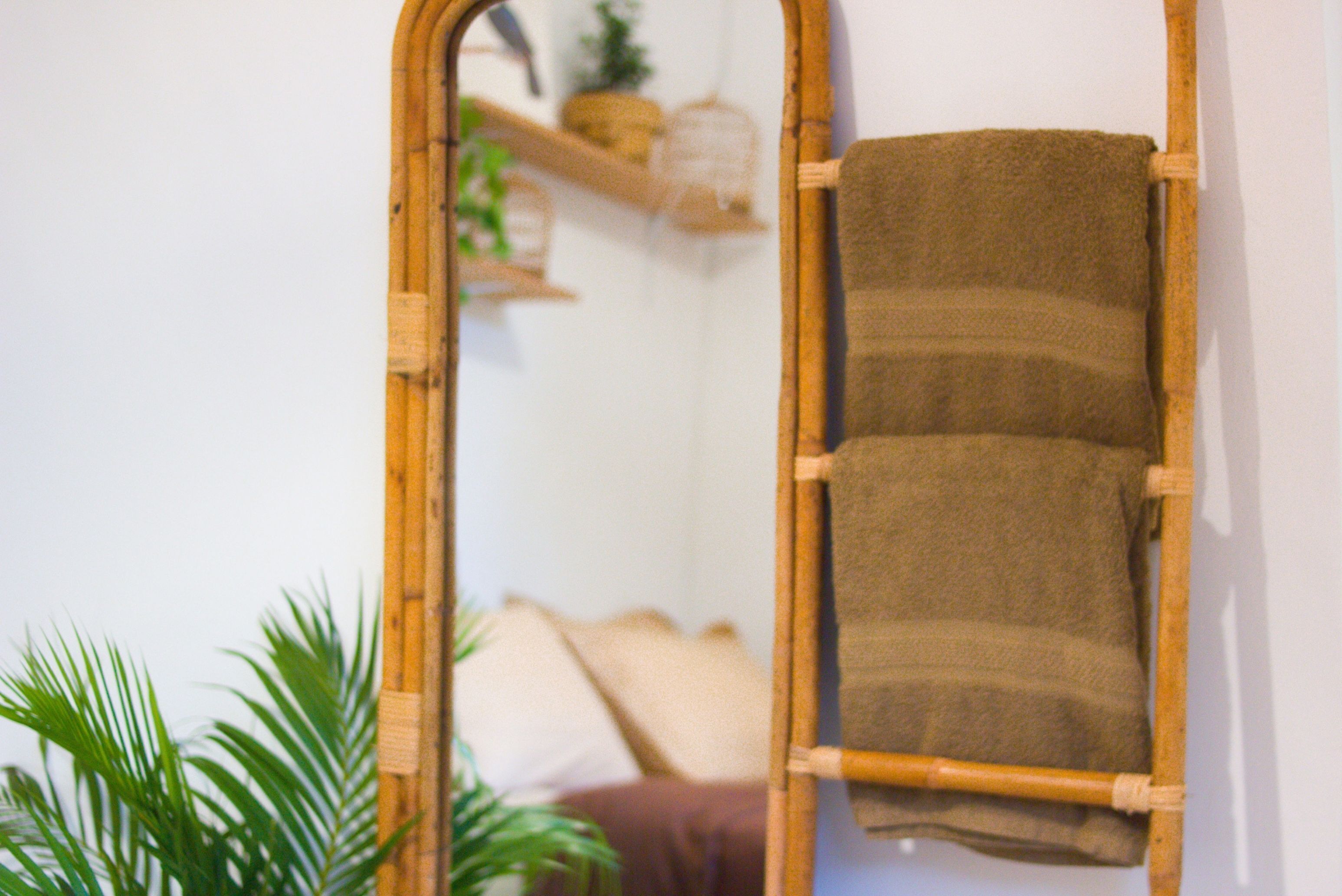 A mirror with a wooden frame reflecting a room with a brown towel hanging on the wall, a plant, and a shelf with decorative items.