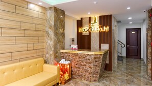 A reception area with a marble counter, a yellow sofa, and a wooden wall. - Moschino HT (Nha Trang)