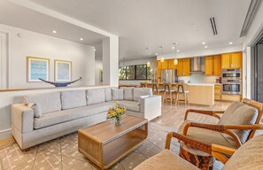 A modern living room with a sofa, coffee table, and chairs, a kitchen with wooden cabinets and appliances, and artwork on the walls.