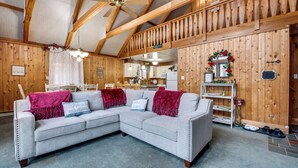A cozy, well-lit room with a large L-shaped sectional sofa, wooden beams on the ceiling, and a kitchen area in the background. - Fire Pit + Community Perks: Gouldsboro Cabin! (Gouldsboro)