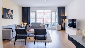 En moderne stue med sofa, to stole, et sofabord, et fjernsyn og en balkon med udsigt til bygninger.